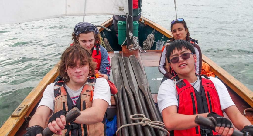 Four people wearing life jackets sit in a boat. Two of them are using oars. 